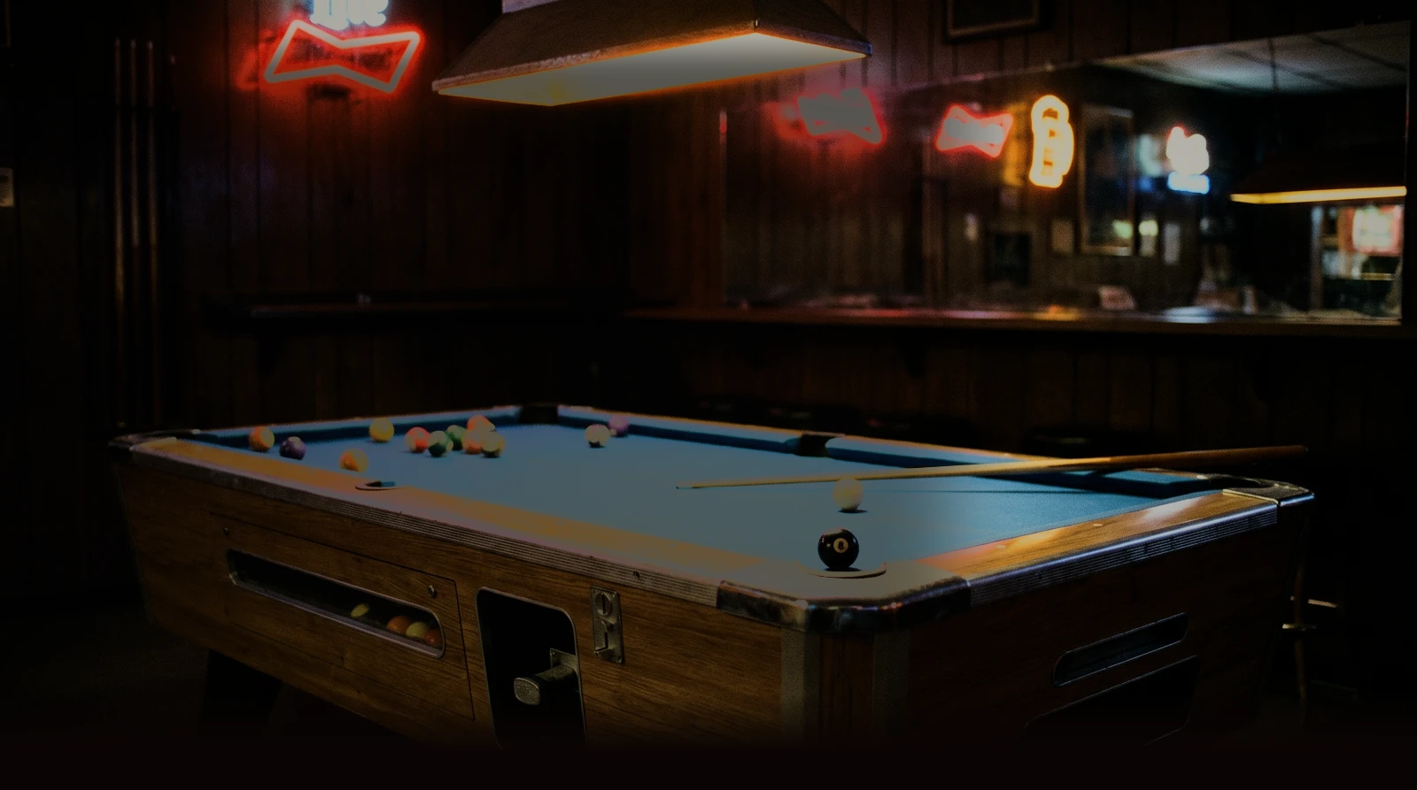 A coin-operated bar pool table lit under its lamp in a neon-signed dive bar on the Social Amusements route
